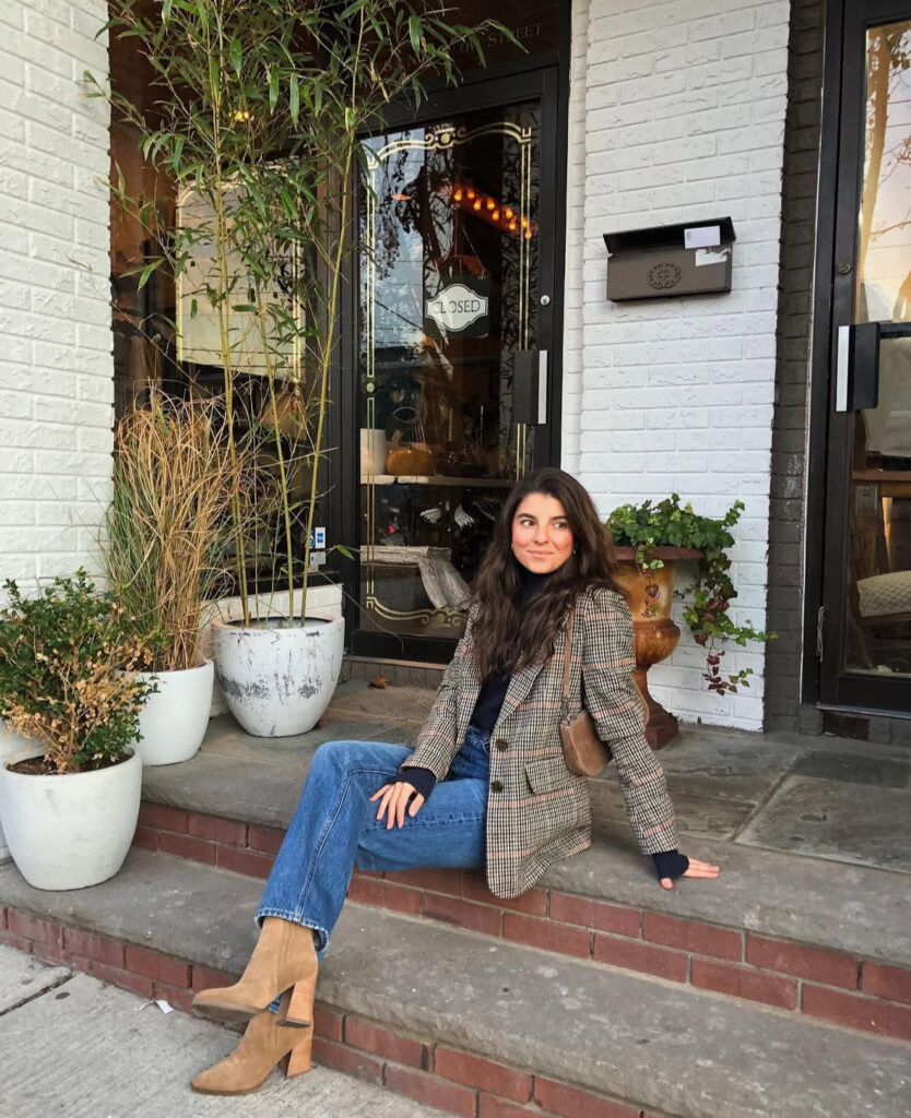 Woman seated on storefront steps in a plaid blazer, navy knit, straight blue jeans, camel block-heel boots, and a small brown shoulder bag.