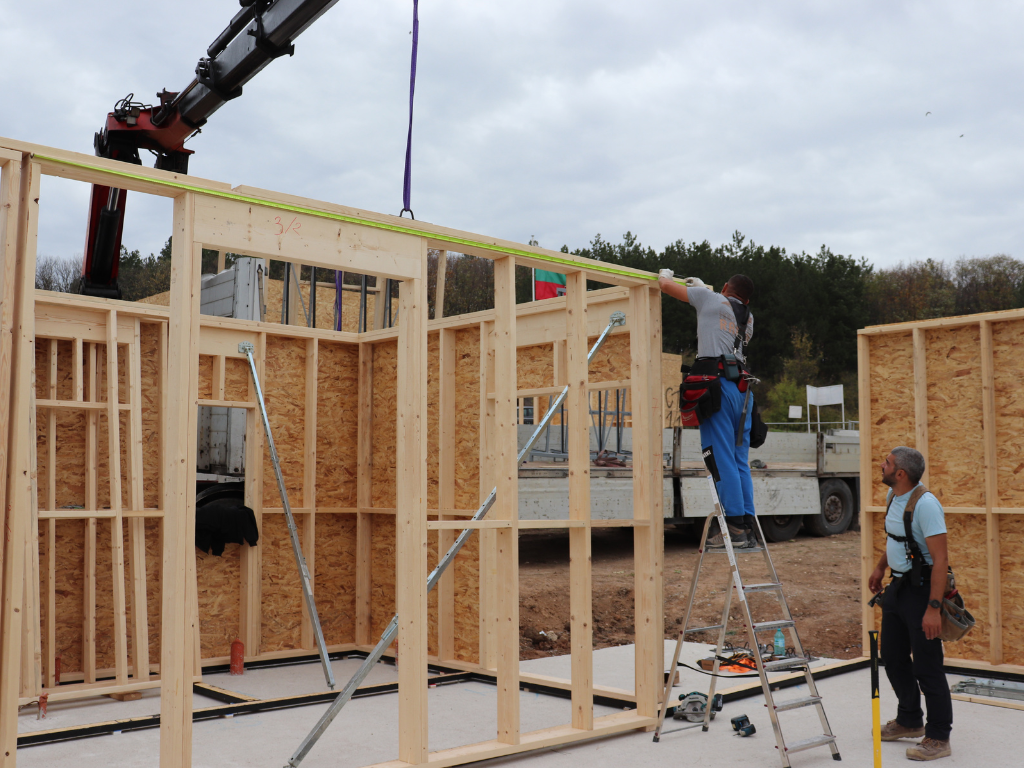 First floor framing with crew setting a header/top plate under a crane, OSB walls going up.