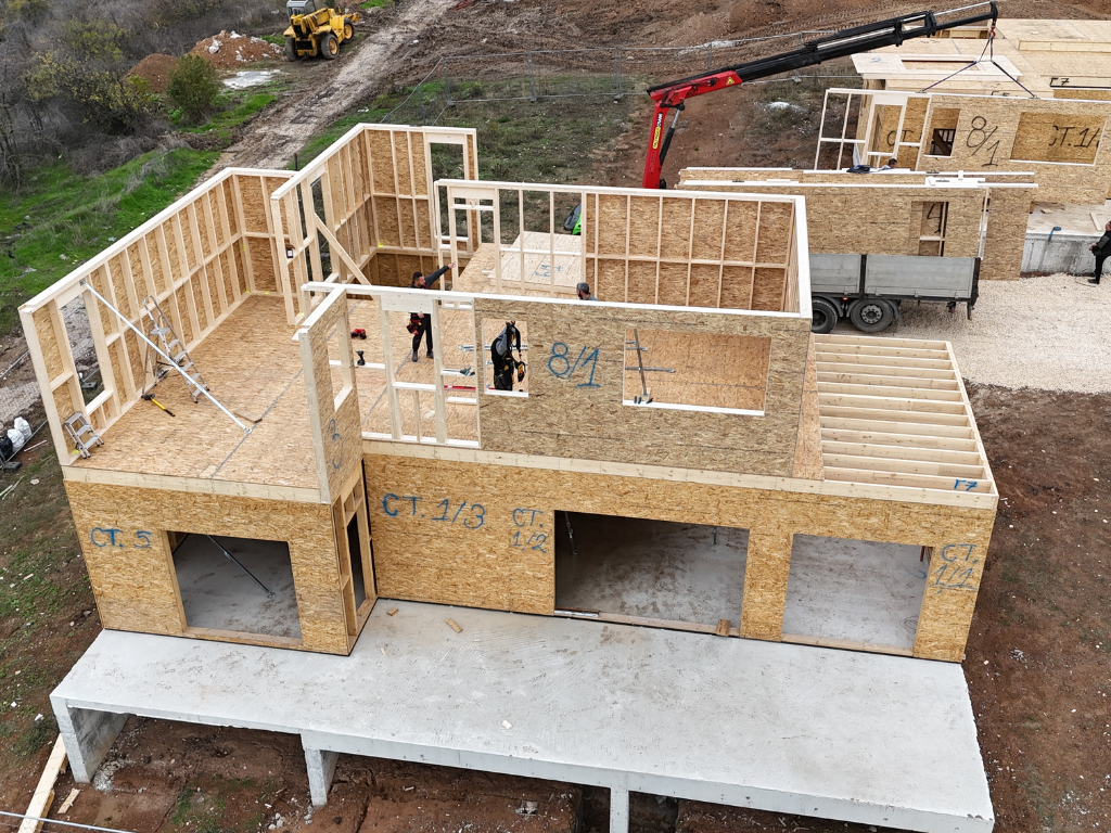 A crane truck unloads prefabricated wooden wall panels beside a partially built two-story house, where workers are installing upper-floor framing on an active construction site.