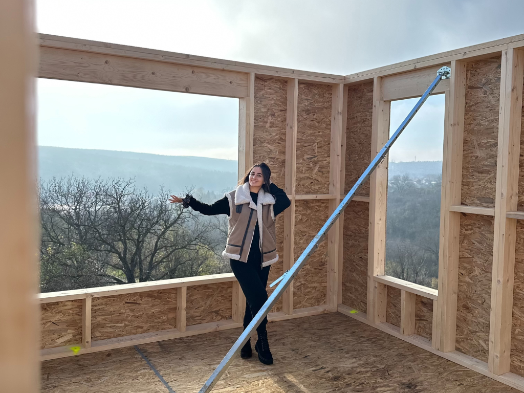 A person stands smiling inside a partially built wooden-framed room with large window openings overlooking a landscape of trees and hills, with a diagonal support brace positioned across the space