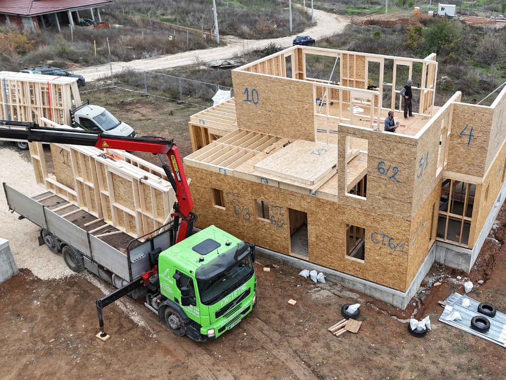 A crane truck unloads prefabricated wooden wall panels beside a partially built two-story house, where workers are installing upper-floor framing on an active construction site.