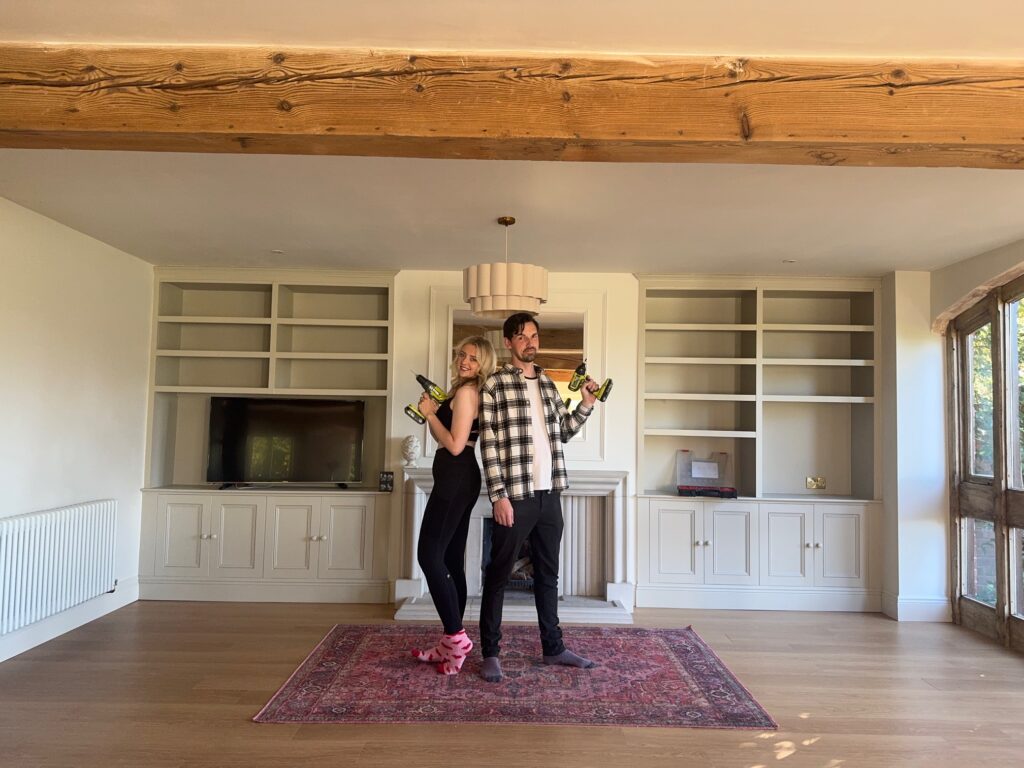 Georgia and Josh posing with drills in their living room with built-in shelving and fireplace, showing DIY barn conversion ideas for cozy spaces.