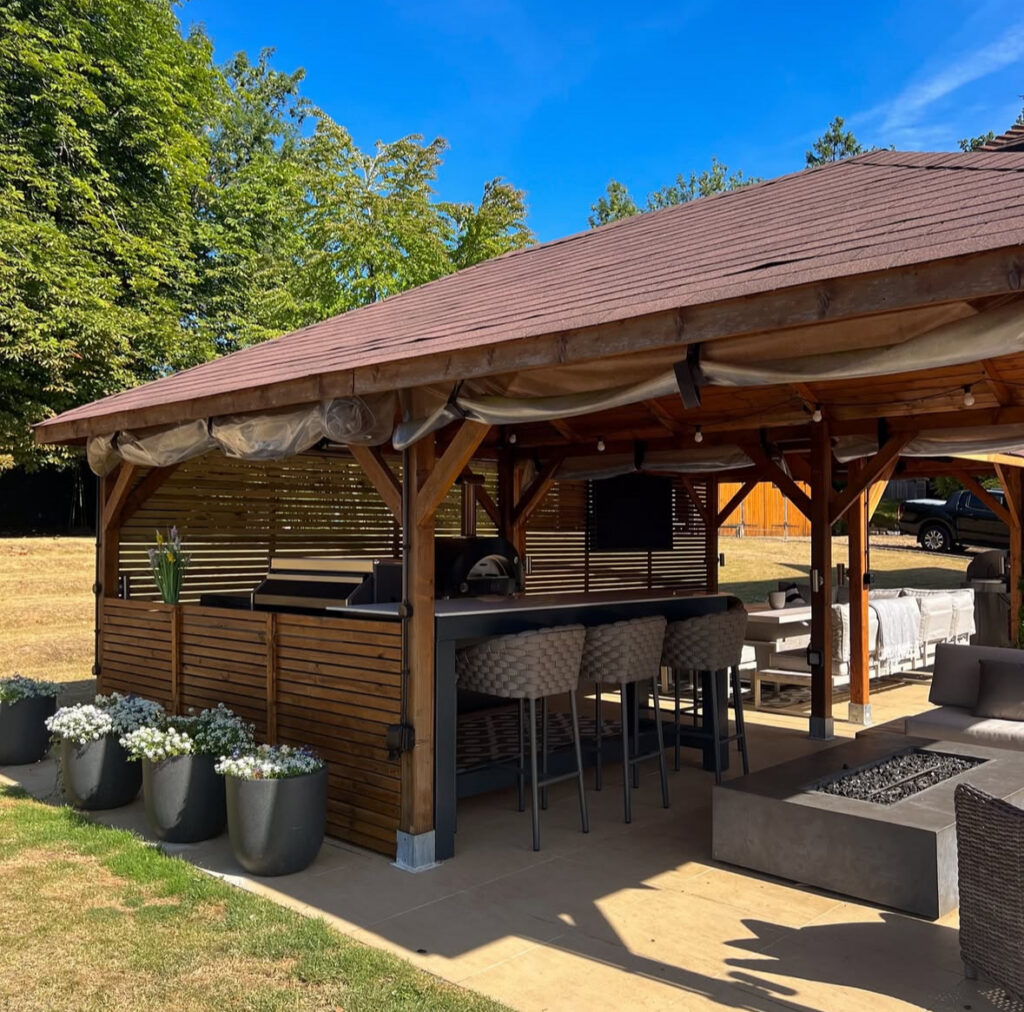 Gabled wood pavilion with bar seating, grill station, and adjacent firepit providing inviting backyard covered outdoor kitchen ideas