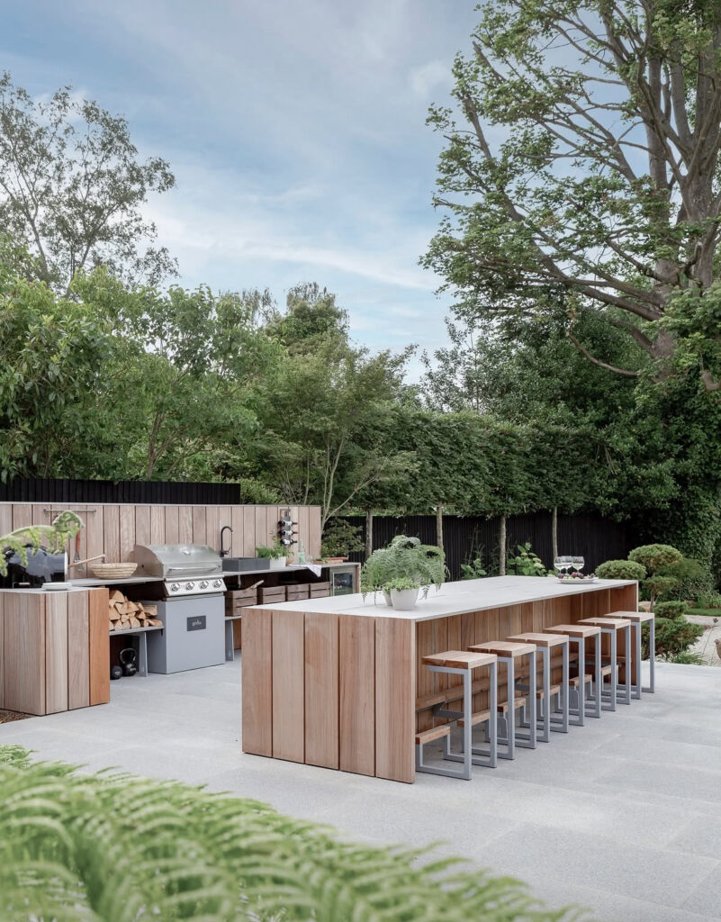 Cedar-wrapped grill wall and long matching dining island defining a generous outdoor kitchen layout in a green garden