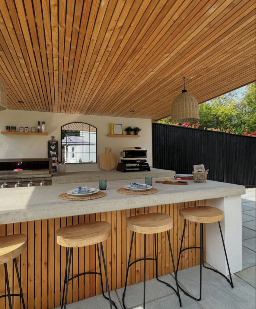 Concrete bar with wood slat ceiling, stools, and open shelving providing relaxed backyard outdoor kitchen ideas.