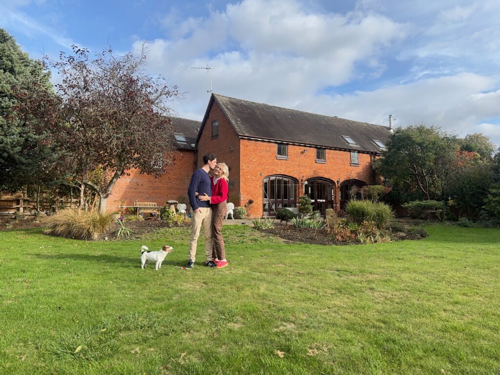 Georgia and Josh standing with their dog in front of their red-brick barn home and garden, a charming snapshot of barn conversion ideas brought to life.