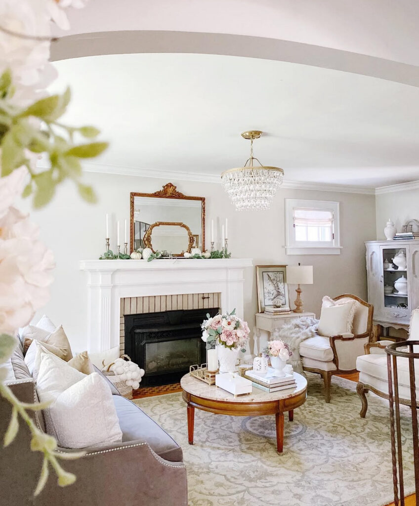 Soft blush florals, vintage mirror, and creamy seating gather around a round coffee table beneath simple french country curtains.