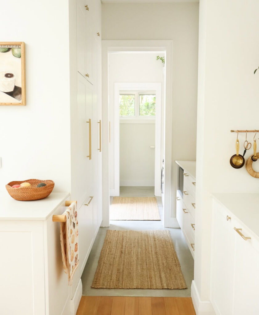 light filled corridor lined with white cabinets and jute runners acts as a narrow walk in pantry connecting spaces