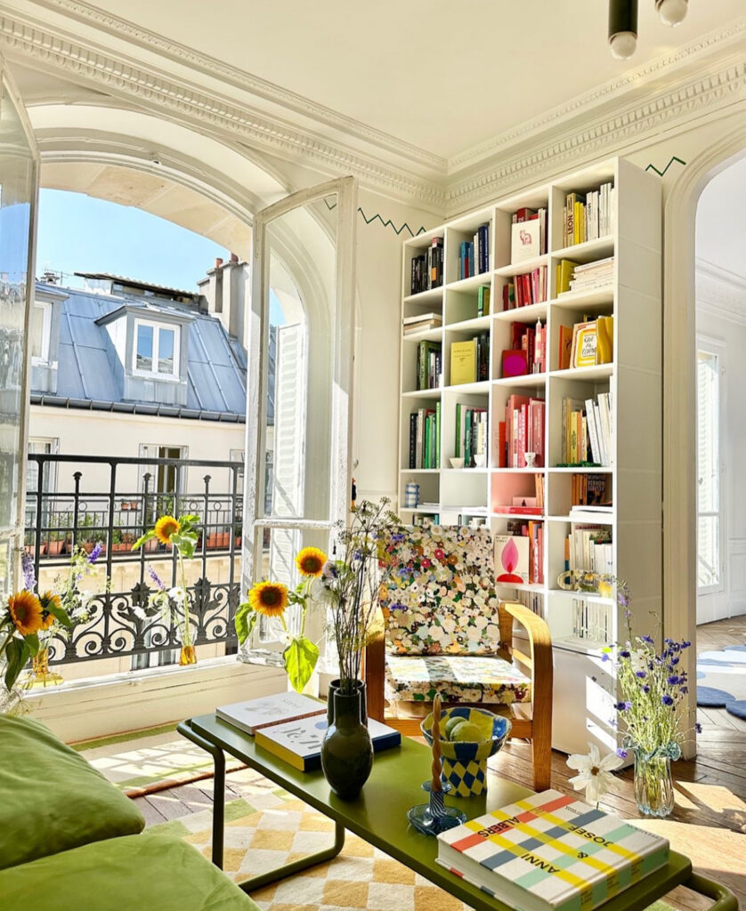 Sunlit Parisian living room with arched windows, colorful bookshelf, and fresh flowers, offering chic European eclectic home decor inspiration.