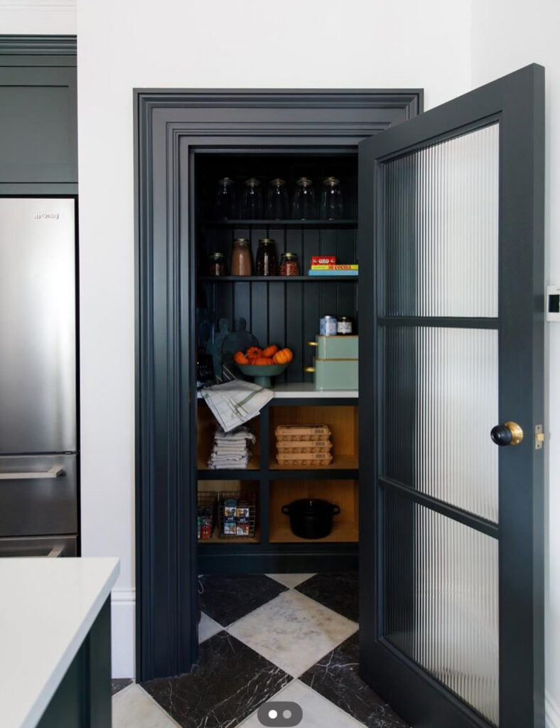 dark beaded board interior behind a ribbed glass door functions as a stylish walk in pantry cabinet with jars and baskets