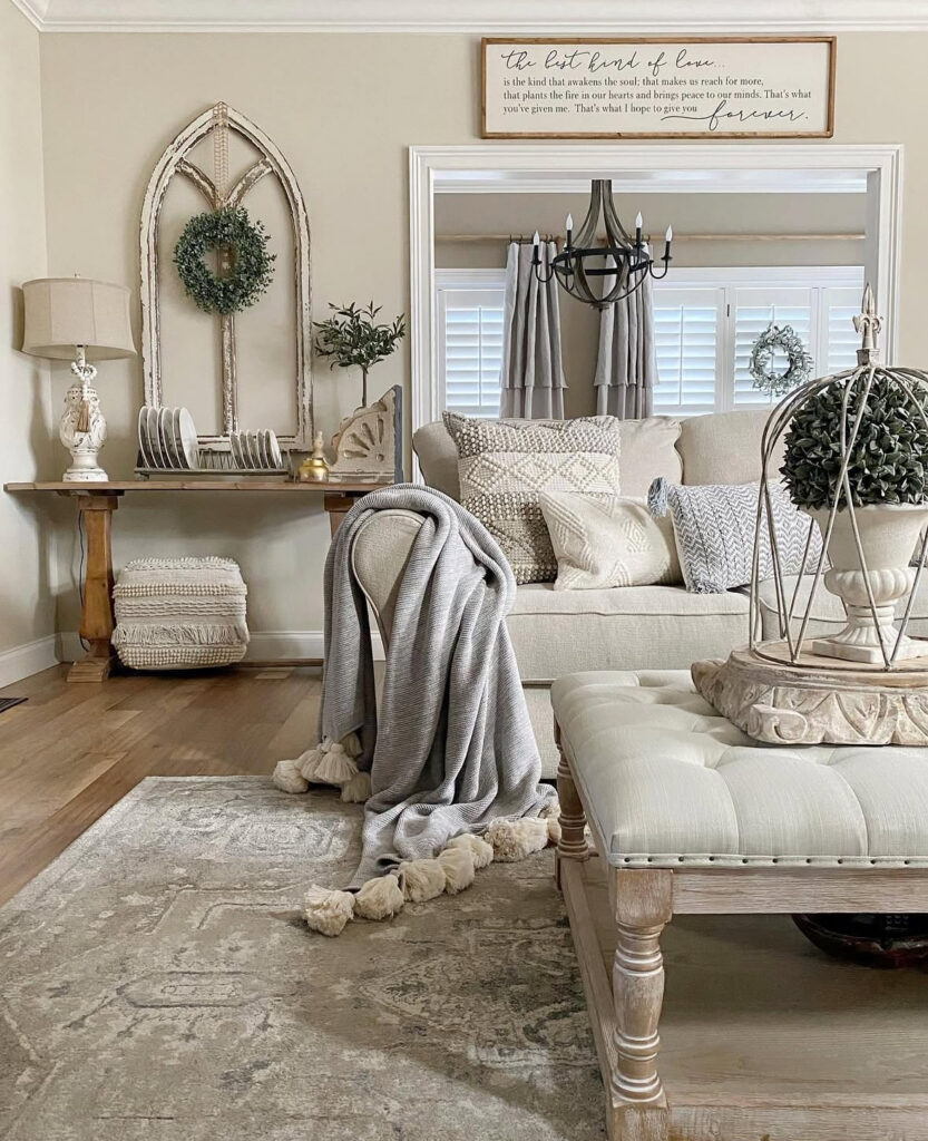 A tufted bench, layered pillows, and long ivory french country curtains frame a warm neutral living room.