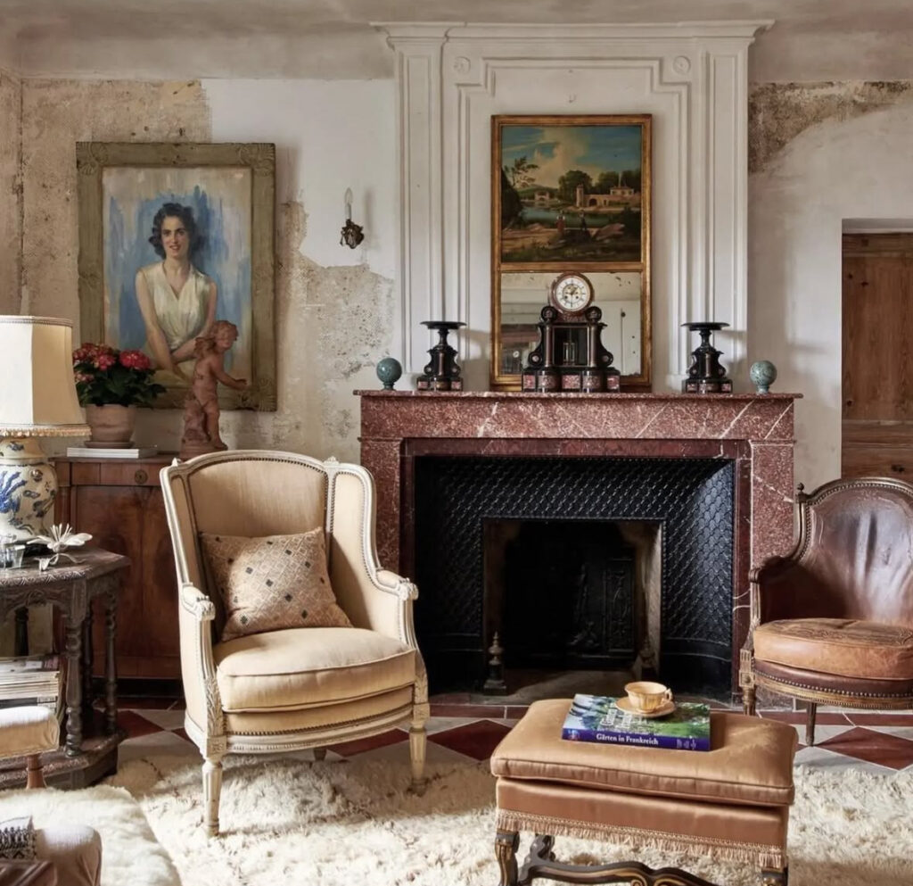 Vintage armchairs and a red marble fireplace in a modern french country living room with layered artwork and timeworn walls.