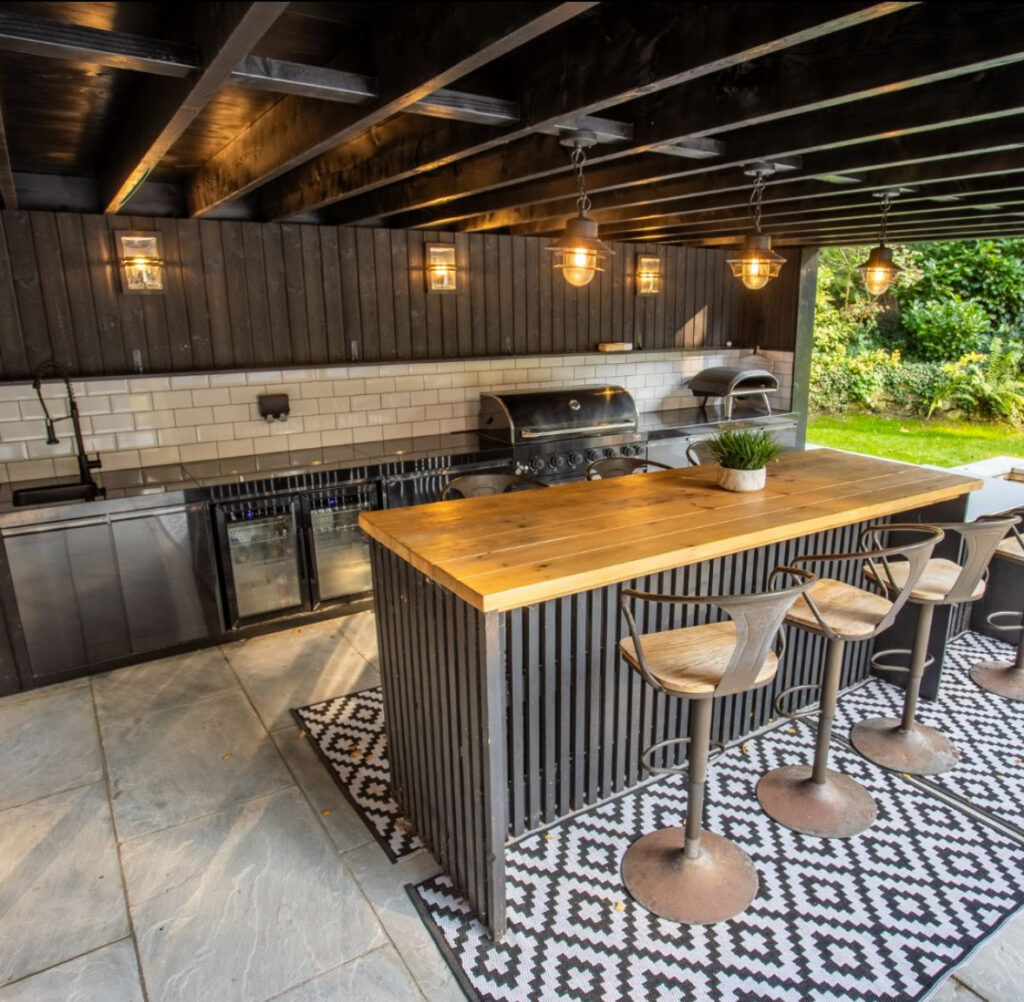 Dark timber bar with pendant lights, patterned rugs, and full-length grill run giving industrial backyard outdoor kitchen ideas