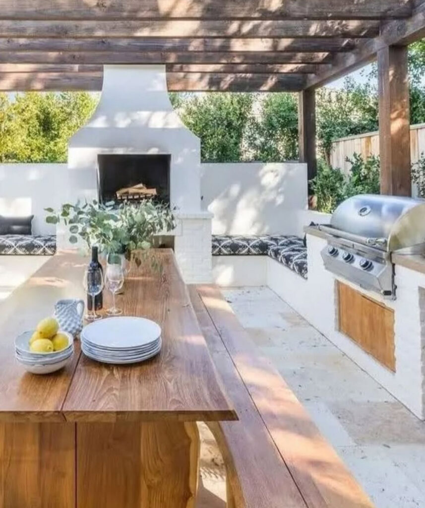 White stucco fireplace, built-in bench seating, and grill around a long table offering charming backyard outdoor kitchen ideas