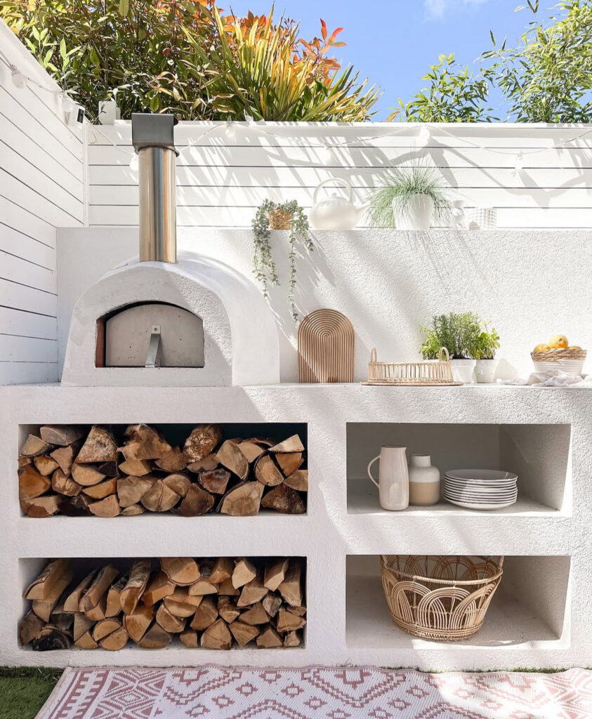 White stucco pizza oven with built-in log storage and open shelves demonstrating a bright cheap simple outdoor kitchen