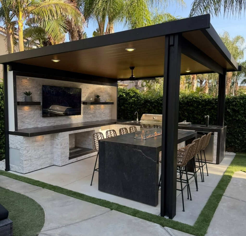 Contemporary pavilion with TV wall, grill, and fire-top dining island showcasing luxe backyard covered outdoor kitchen ideas