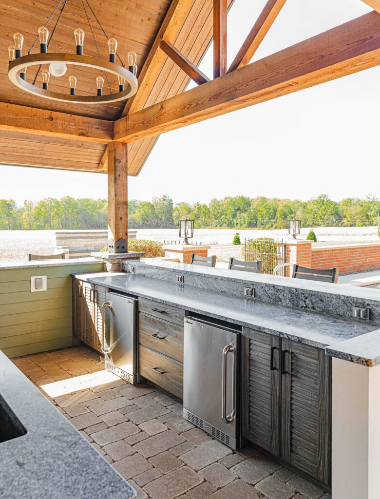 Covered bar with undercounter fridges and stone countertop beneath a timber pavilion illustrating a practical outdoor kitchen layout.