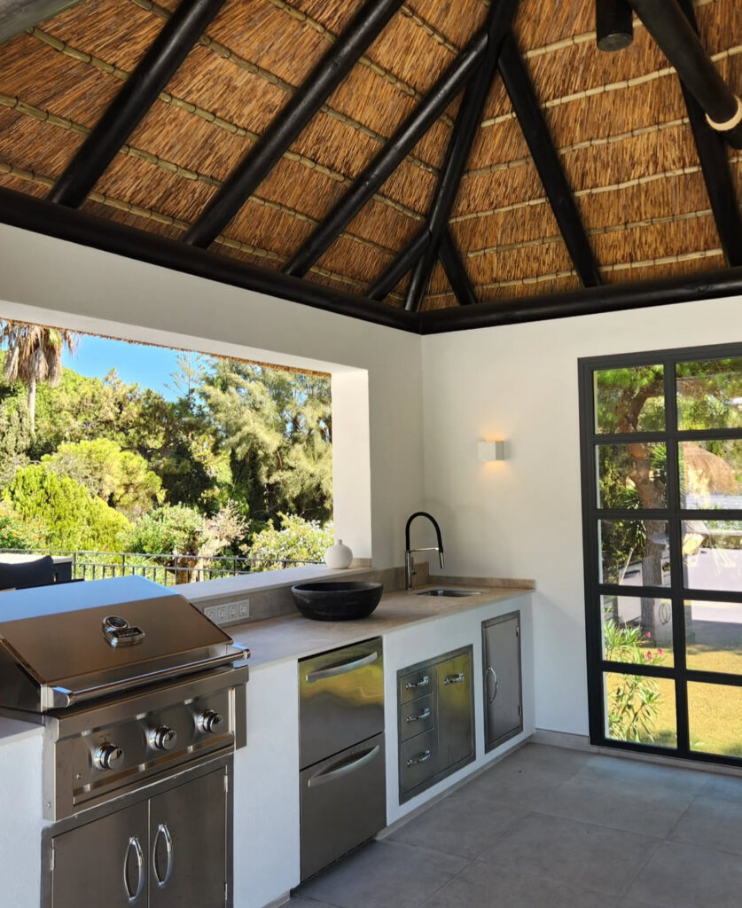 Thatched-roof structure with stainless grill, sink, and counter arranged in an airy outdoor kitchen layout