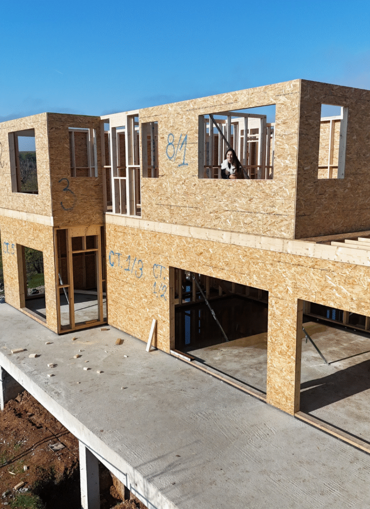 Partially constructed two-story house with OSB-covered exterior walls and visible framing, built on a concrete platform; a person looks out through an upper-floor window opening on a clear, sunny day