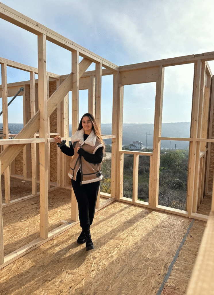 A person stands smiling inside a partially framed house, holding onto a wooden stud, with large window openings behind them showing a bright landscape of hills and scattered houses
