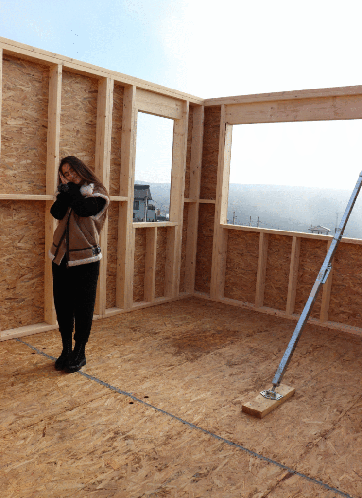 A person poses warmly inside a partially built wooden room with exposed framing and large window openings overlooking distant houses and a misty landscape