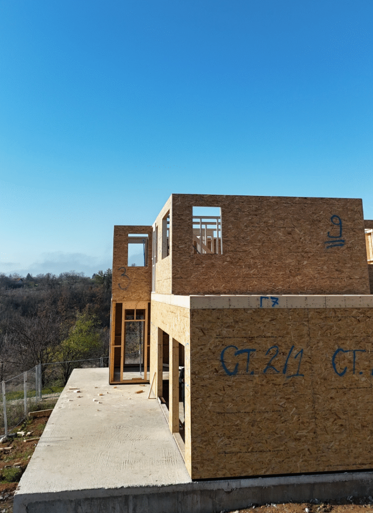 Two-story wooden-framed house under construction on a concrete slab, with OSB exterior walls, labeled sections, and window openings visible against a clear blue sky