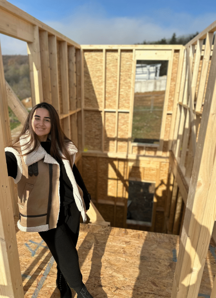 A person stands smiling inside an unfinished, wooden-framed upper floor of a house, with an open drop to the lower level behind them and outdoor scenery visible through the framing