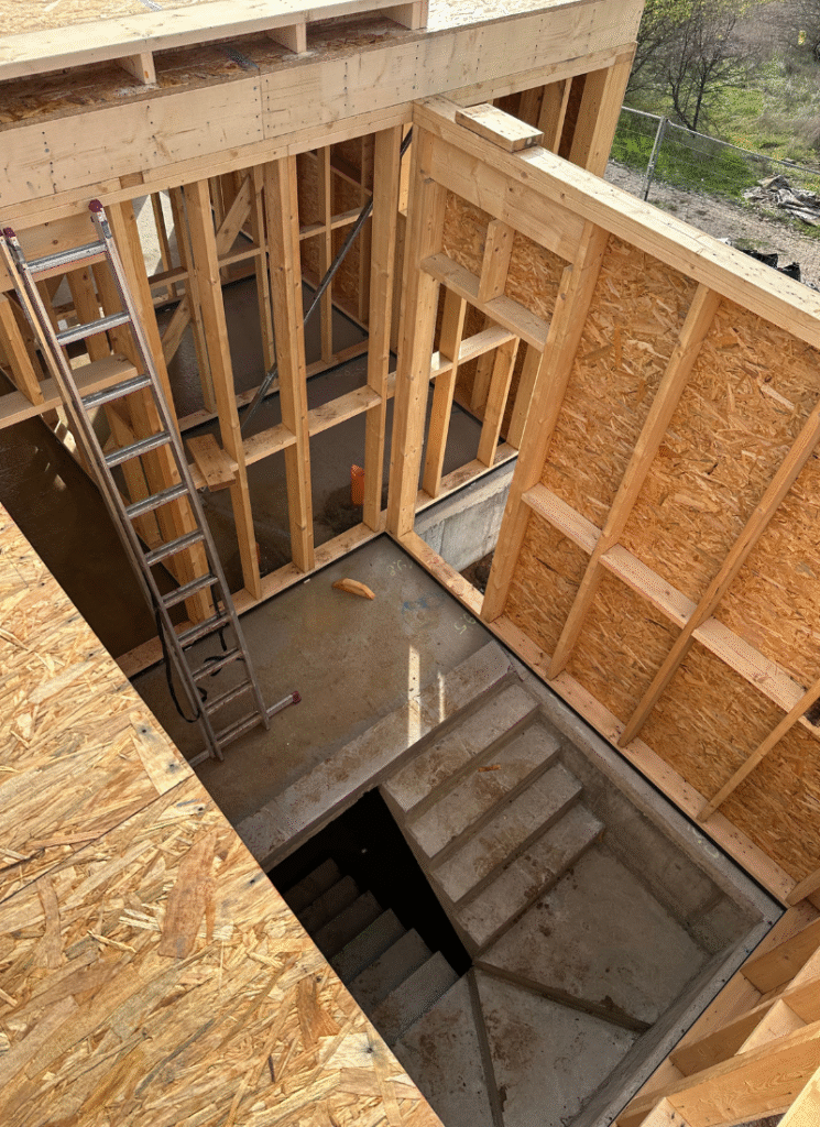 View from an upper level of a house under construction, showing exposed wooden framing, a metal ladder, and a concrete stairwell leading down to the lower floor