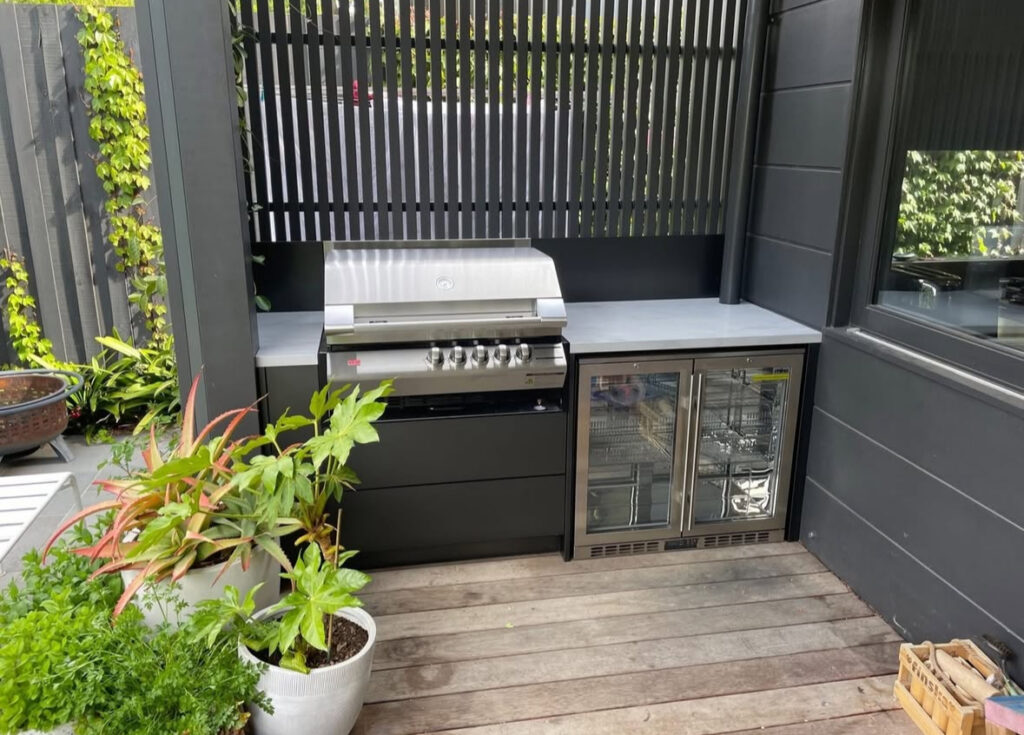 Dark cabinetry, stainless grill, and glass-front fridge fitted into a tight deck corner as a sleek small outdoor kitchen