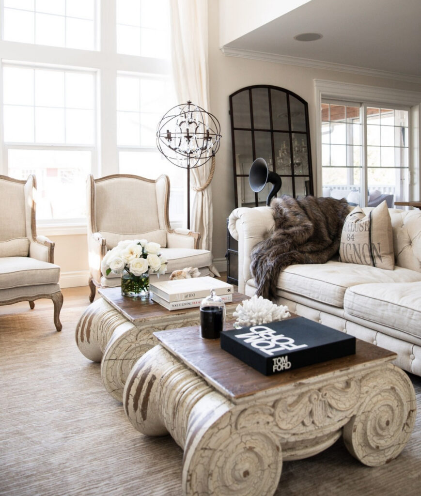 Grand wingback chairs, tufted sofa, and chunky carved coffee tables define a rustic french country living room bathed in natural light.