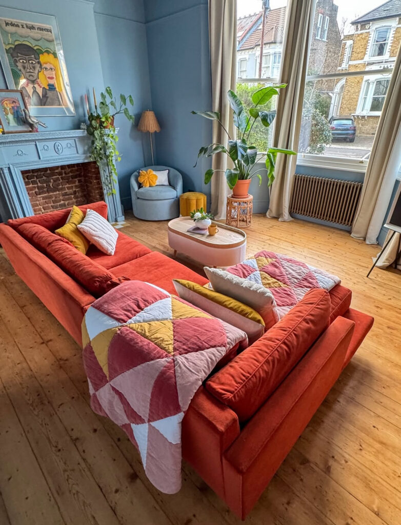 Colorful 70s decor living room where a vivid orange sofa contrasts with soft blue walls, big windows and plants.