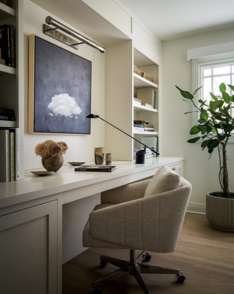 Cream built-in home office wall with long white desk, taupe swivel chair, shelves of books and a framed cloud painting lit by a picture light.