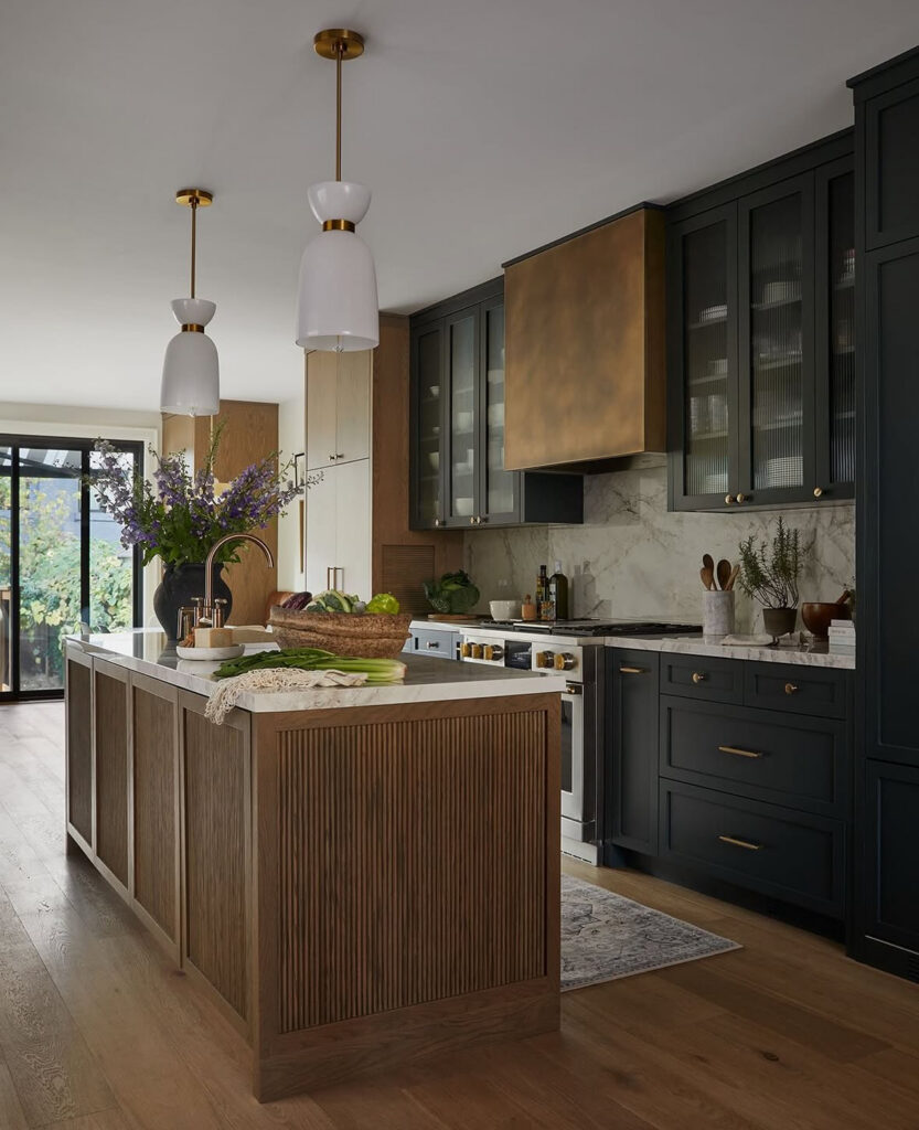 Luxe loft kitchen with dark custom cabinetry, bronze hood, long fluted wood island and marble walls designed by @dartstudioinc.