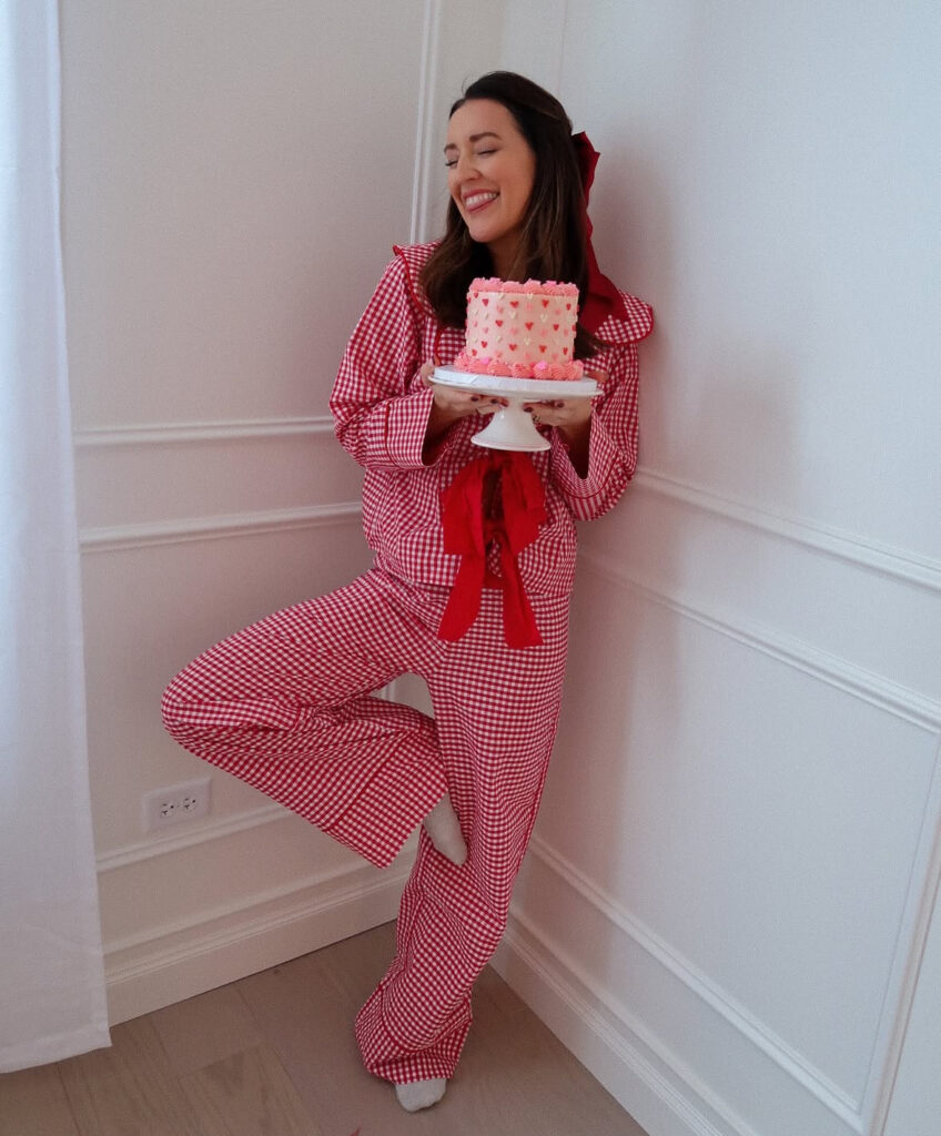 Woman in cozy pink pajama-style Galentine’s outfit holding a heart-shaped cake indoors