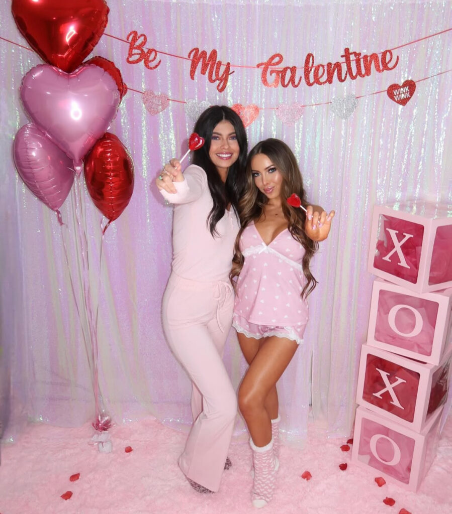 Women in blush and pink Galentine’s outfits posing in front of a Valentine-themed party backdrop