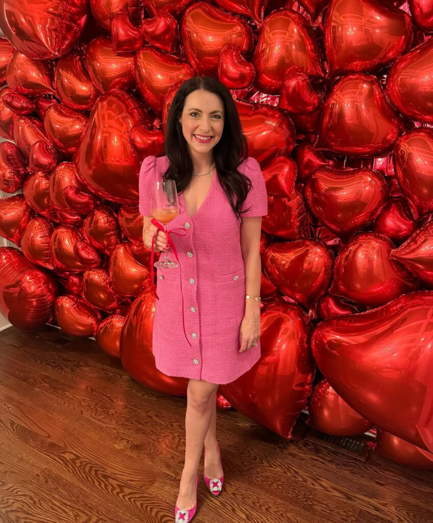 Woman wearing a pink Galentine’s dress posing in front of a red heart balloon wall at a party