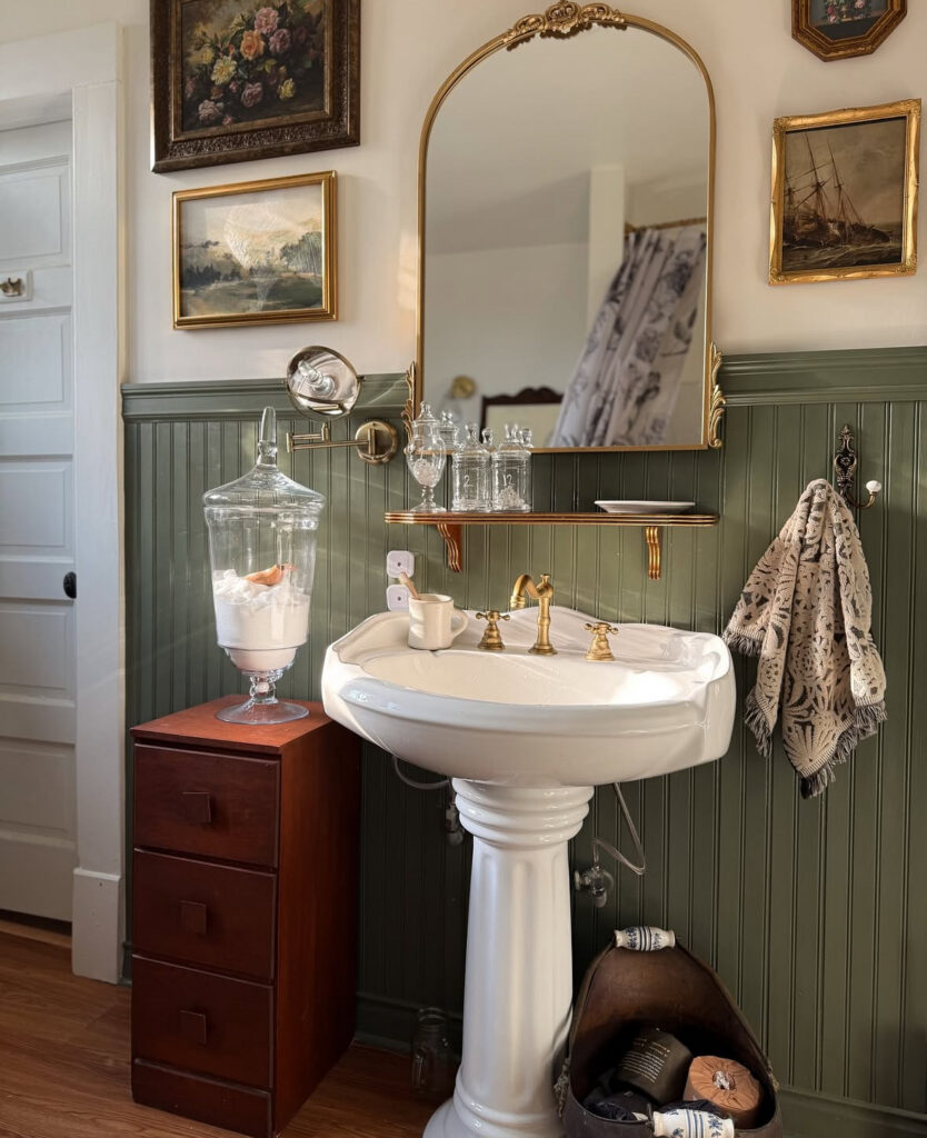Pedestal sink against green paneling topped with an arched gold vintage bathroom mirror and framed art.