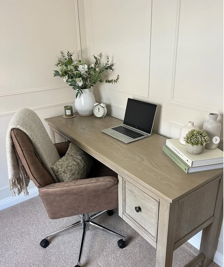 Styled desk against paneled walls with a vase of flowers, a small clock, and a cozy chair with a throw.