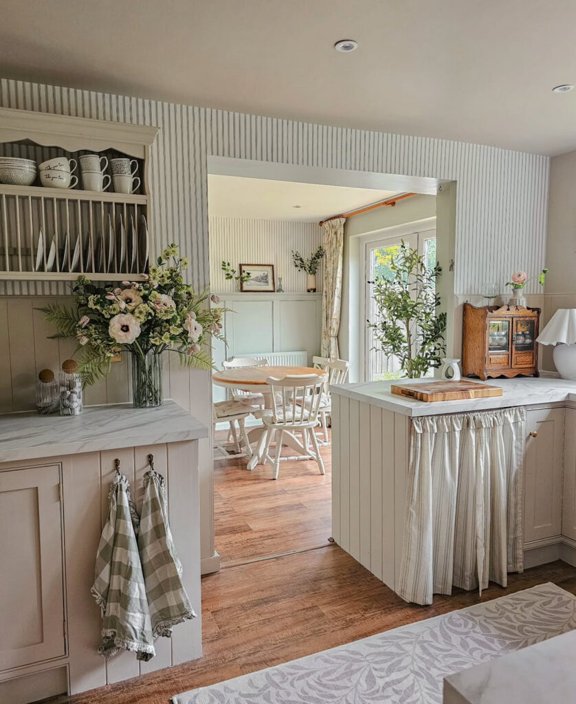 Soft modern country kitchen with vertical panelling, skirted sink, open shelving and a cosy dining view created over time by @pagesofemma.