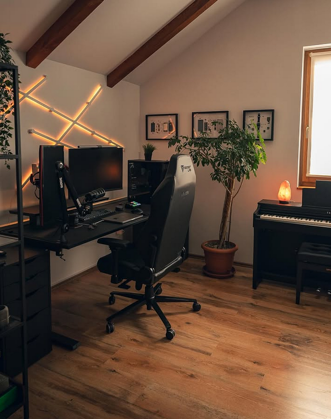 Cozy loft workspace with warm wood floors, LED wall lights behind a dual-monitor desk setup, a large chair, framed art, and a piano with a small glowing lamp.