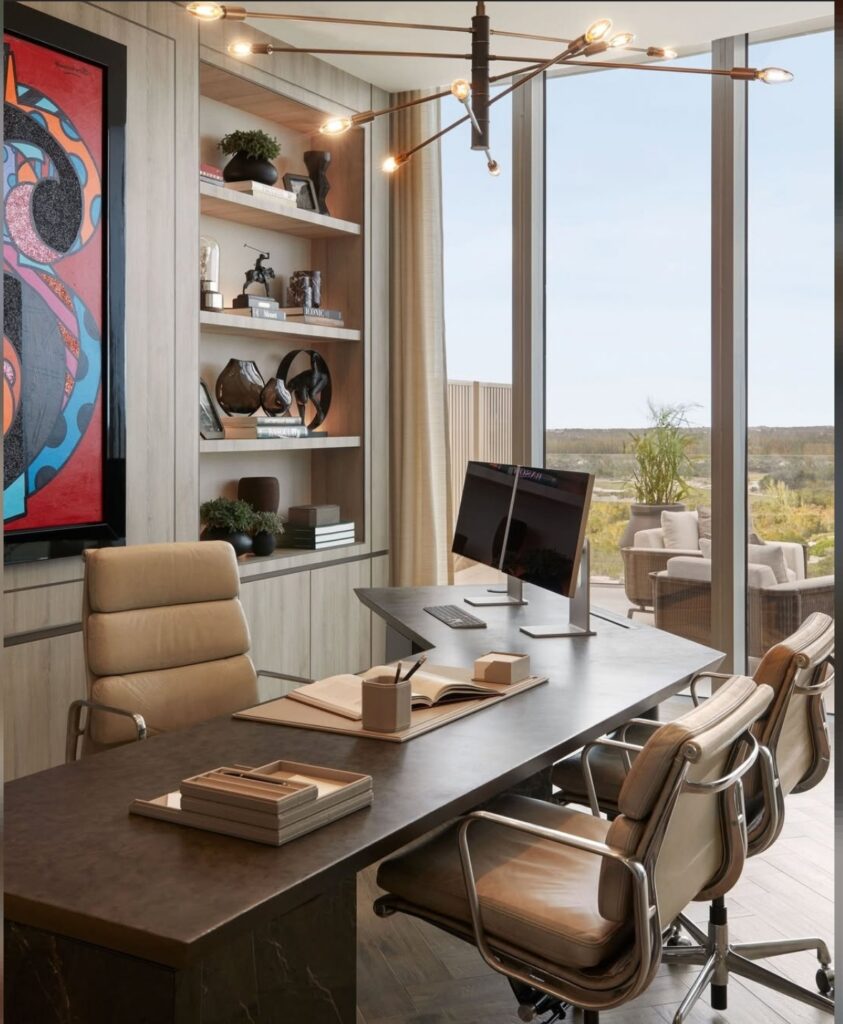Luxury modern home office with dark stone desk, tan leather swivel chairs, large colorful abstract art, open shelving and full height windows to a terrace view.