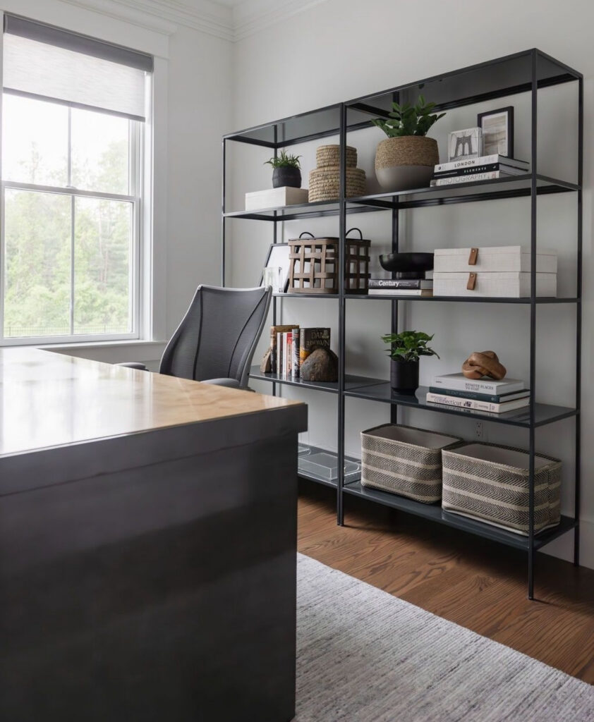 Modern masculine office with black metal shelving styled with baskets and plants, a wood-topped desk, ergonomic office chair and large window