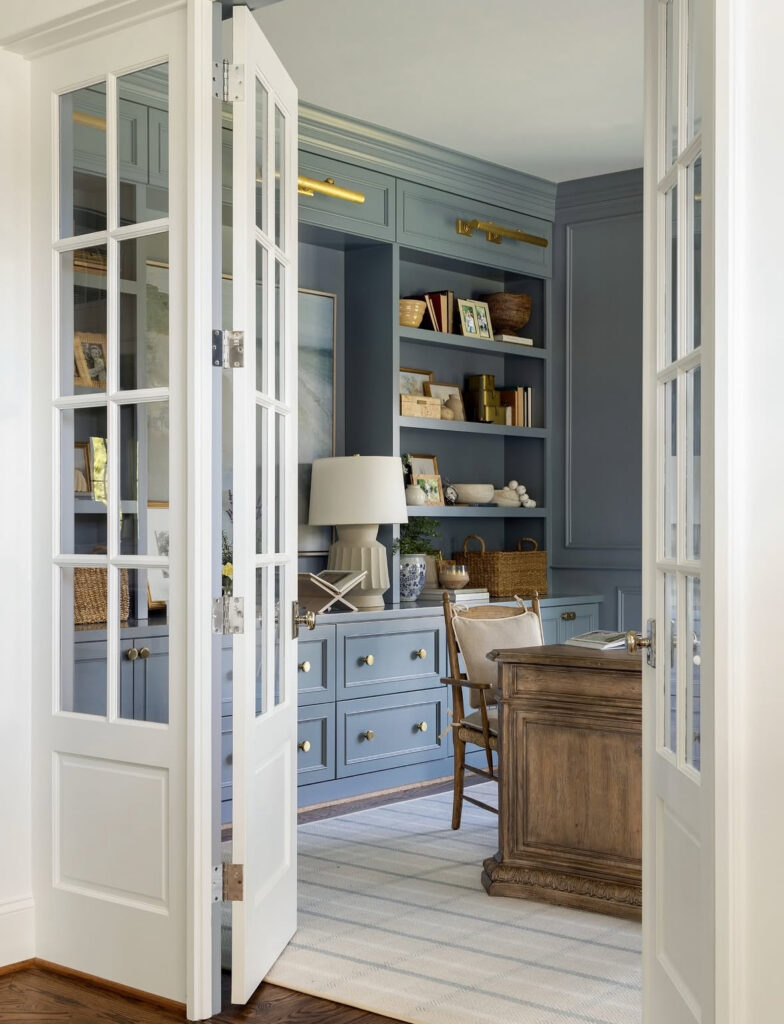 Masculine home office with blue built-in cabinets, glass French doors, brass picture lights and a traditional wood desk on a pale striped rug