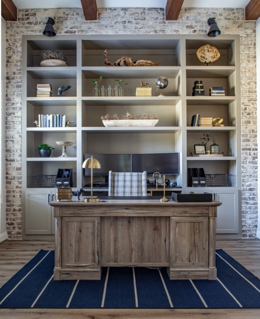 Masculine home office with exposed brick wall, taupe built-in shelves, rustic wood desk and a navy striped rug grounding the space