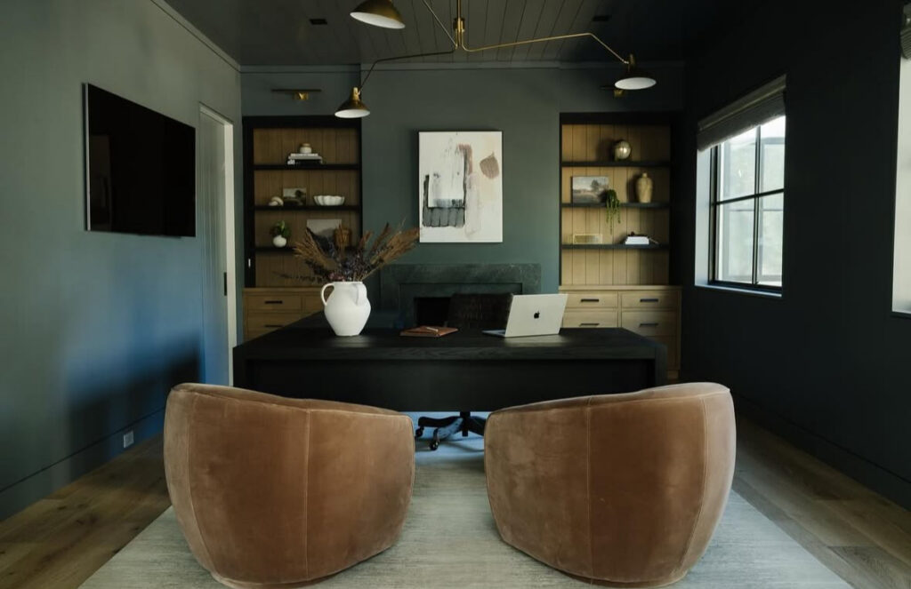 Modern masculine office with charcoal walls, black desk, light wood shelves, two tan swivel chairs and a sculptural brass ceiling light