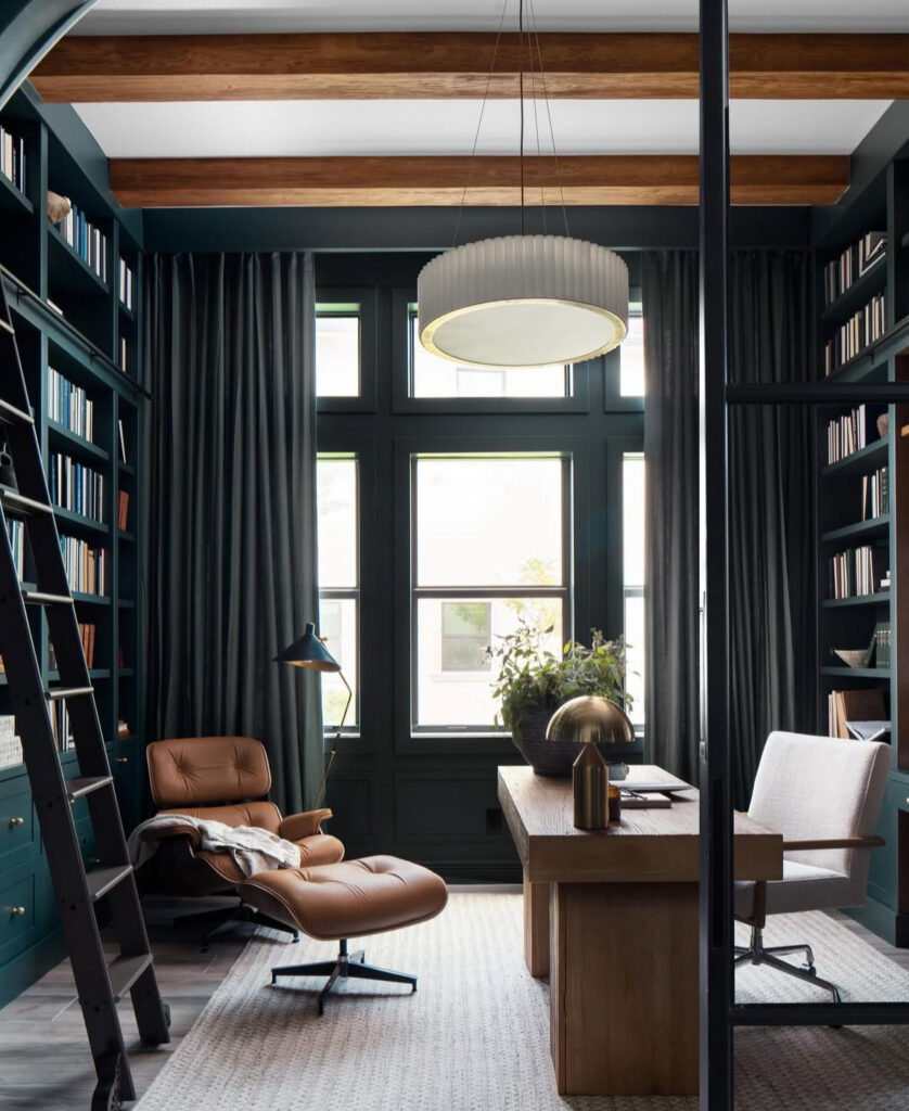 Moody masculine study with deep green built-ins, rolling library ladder, wood ceiling beams, tan leather lounge chair and a chunky wood desk in front of tall windows