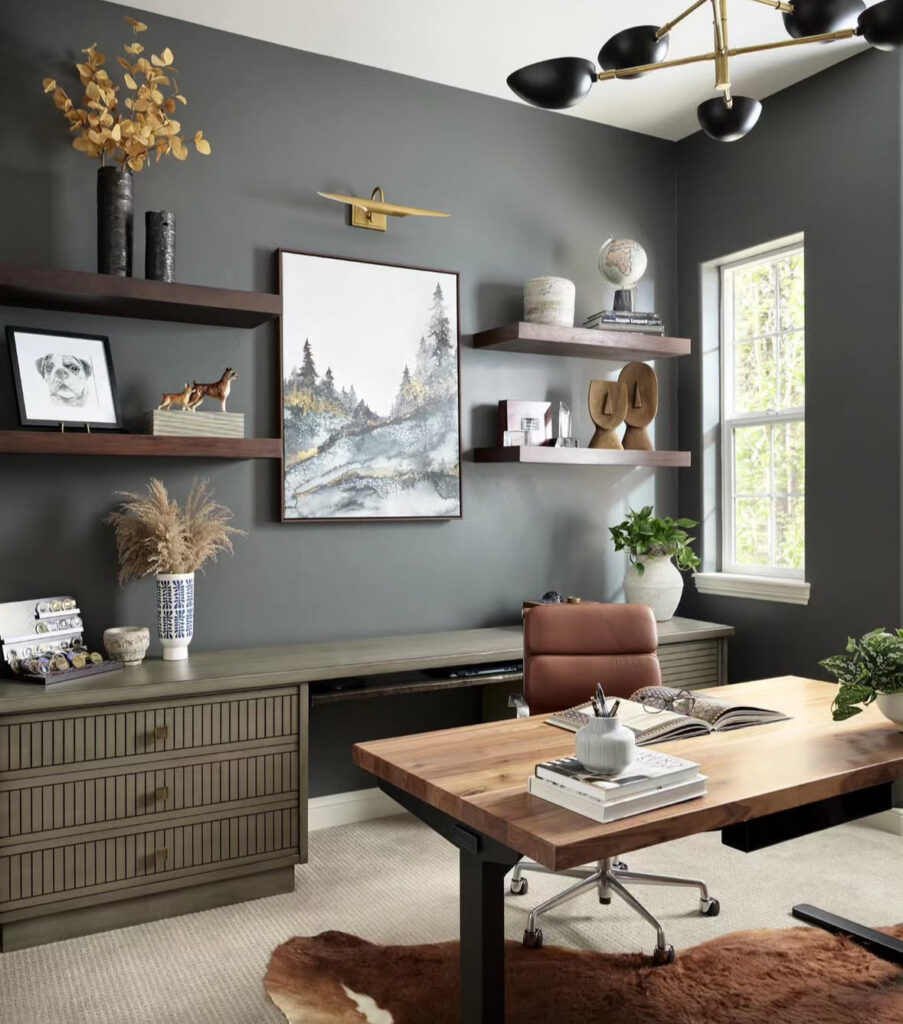 Masculine home office with deep grey walls, wood desk, floating wood shelves, landscape artwork with picture light and a cognac leather chair on a hide rug