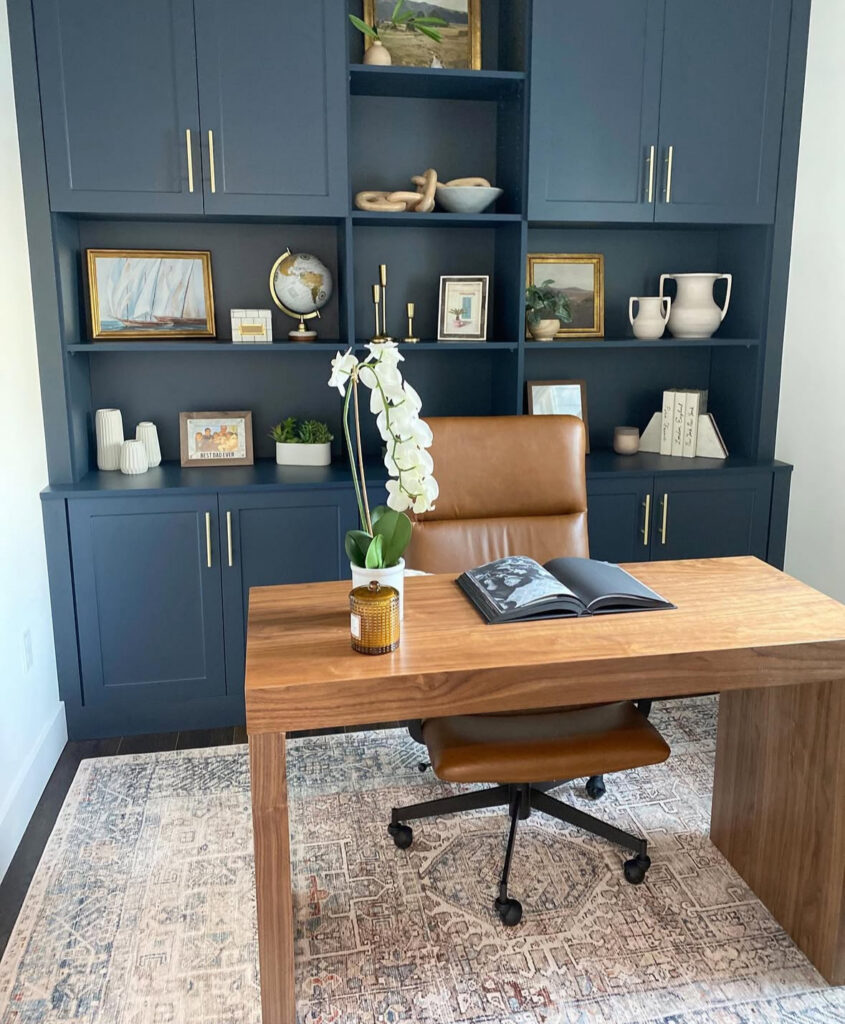Masculine office with navy built-in cabinets, brass hardware, minimalist wood desk, caramel leather office chair and a patterned rug