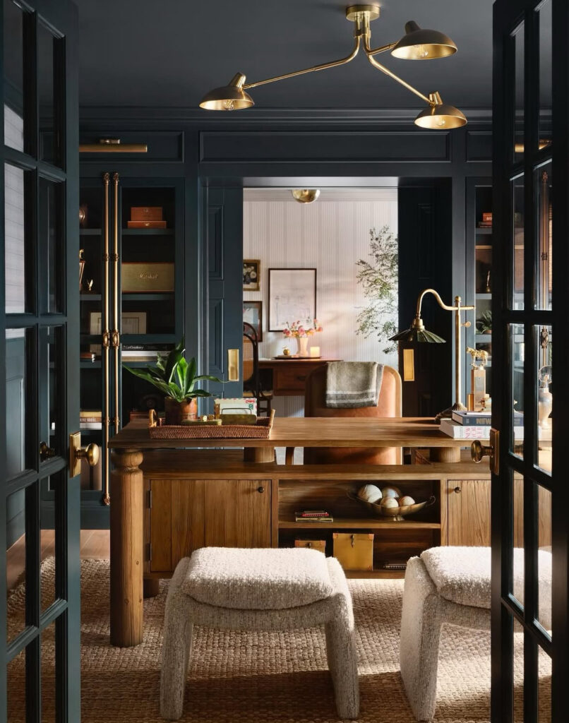 Handsome navy paneled home office with wood desk, leather desk chair, brass lighting, jute rug and two cream boucle stools in front of the desk