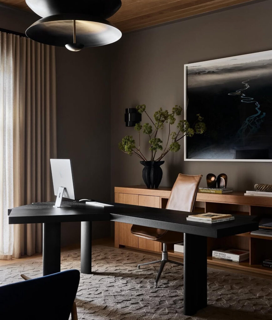 Moody masculine office with warm taupe walls, a sculptural black desk, tan leather desk chair, wood credenza and oversized black pendant light